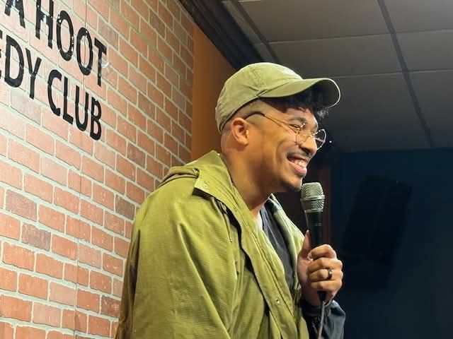 Jeremiah performing with a microphone in front of a brick comedy club wall